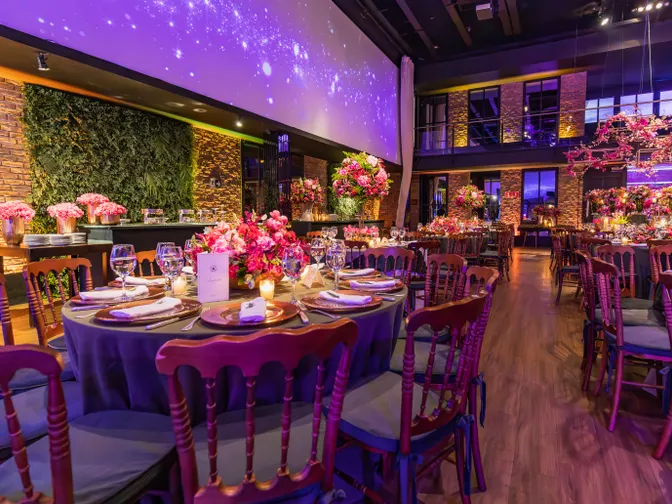 A wedding reception room lit in purple, with round tables dressed in pink linen, tall pink floral centrepieces and gold chairs.