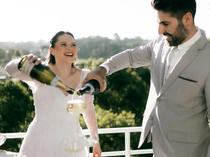 A laughing bride and groom pouring two bottles of champagne into a tower of glasses on a sunlit balcony.
