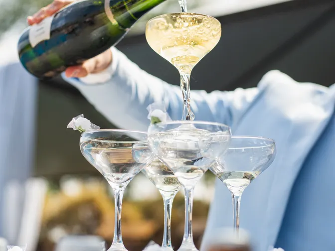 A champagne tower of stacked coupe glasses being filled at an outdoor wedding, with guests and greenery behind it.