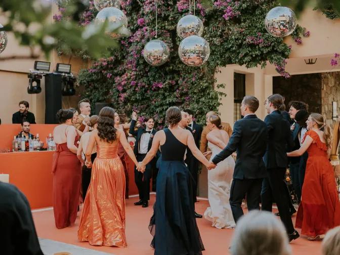 Wedding guests in orange and navy formalwear holding hands in a circle on an outdoor dance floor, under disco balls hung in front of a wall of pink bougainvillea.