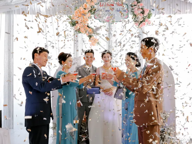 Newlyweds raising a toast with their parents and family under a flower-decorated canopy as confetti falls.