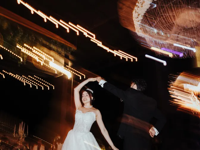 A bride and groom dancing closely under strings of warm fairy lights at an outdoor evening reception.