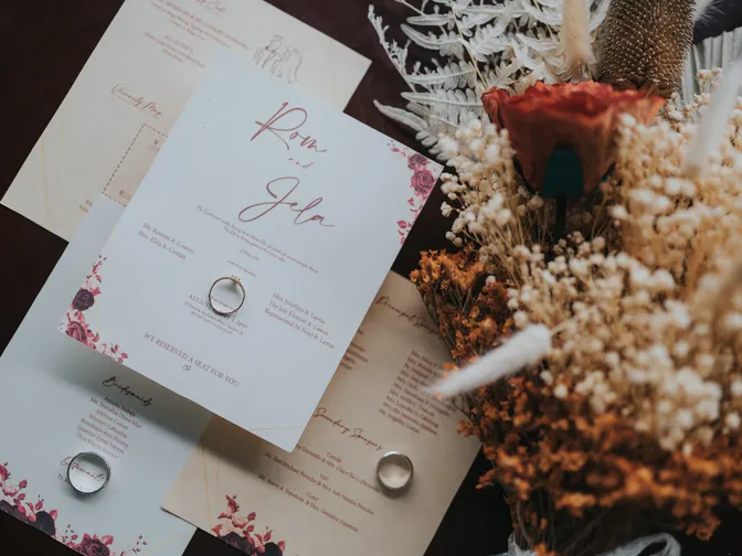 A wedding invitation suite printed in deep red on white card, laid out on a dark table with two rings and a bouquet of dried orange flowers.