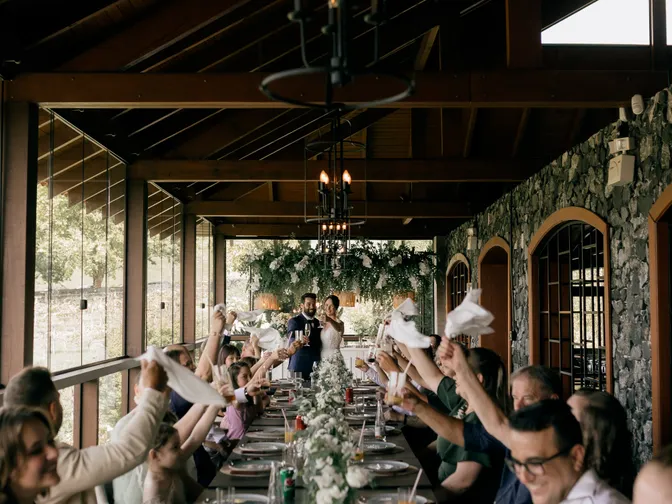 Wedding guests seated along a long table under a wooden roof raising their glasses towards the couple at the far end.