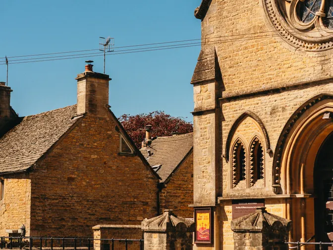 A honey-coloured stone church with an arched doorway and a red parish noticeboard, standing beside an old cottage under a clear blue sky.