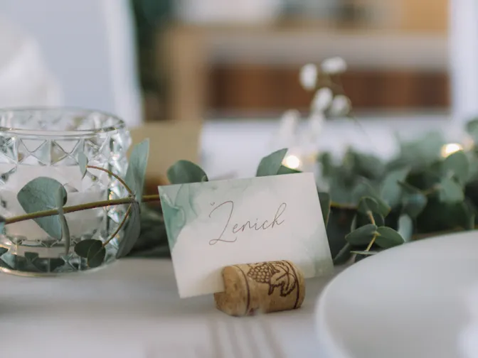 A handwritten place card propped on a wine cork at an elegant wedding table set with eucalyptus and glassware.