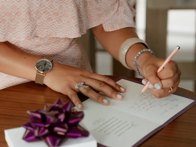 A woman in a pink dress handwriting inside a card on a wooden table, a purple gift bow beside her.